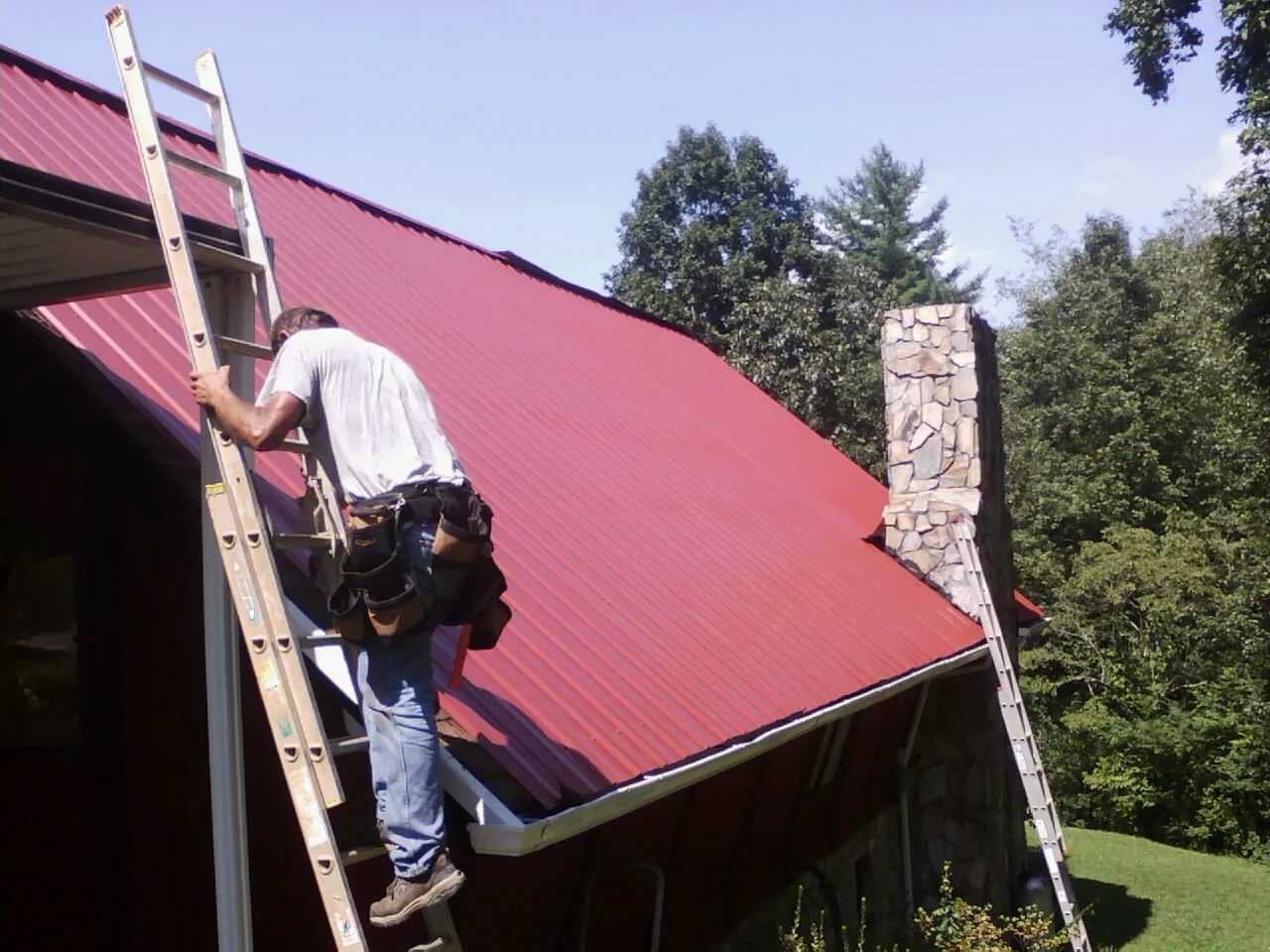 Roofer on a ladder beside a red metal roof and stone chimney — Soffit & Fascia Repair by Roofing Brothers in Tatum